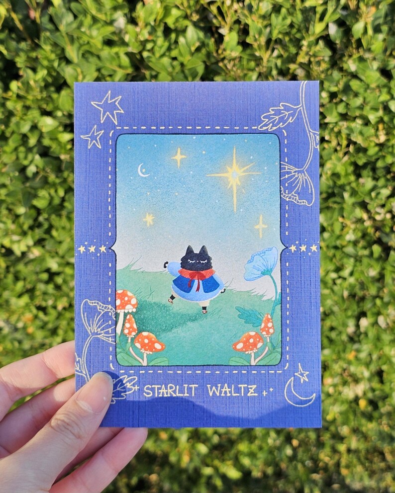 May include: A blue greeting card with a whimsical illustration of a black cat in a blue coat dancing in a field with mushrooms and flowers. The card has the text "Starlit Waltz".