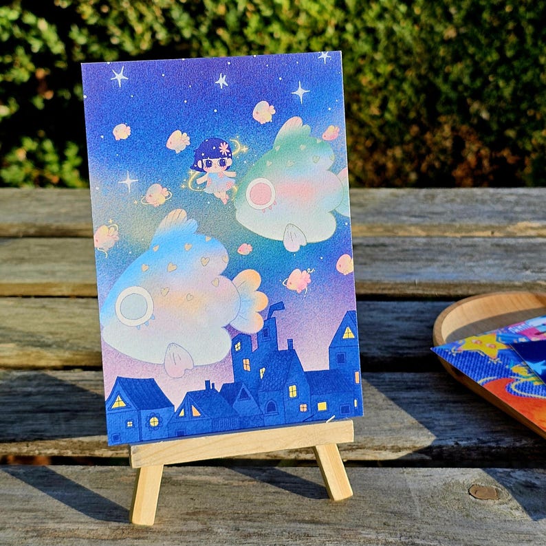 May include: A whimsical illustration on a card, featuring a night sky with stars and floating fish-like creatures. A small figure with a floral headpiece floats among the fish. The bottom of the card depicts a town with blue rooftops and lit windows.