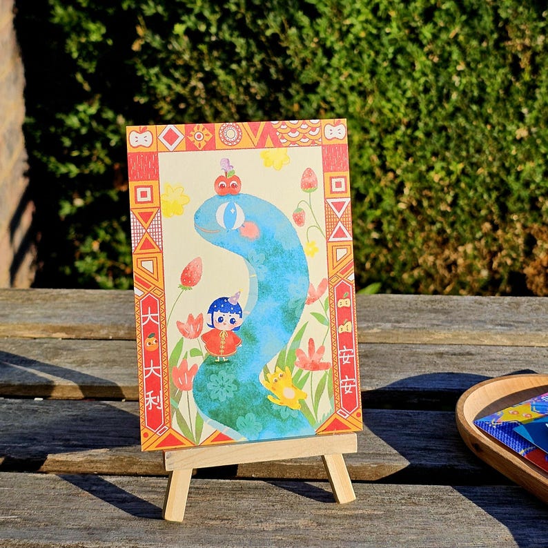May include: A colorful illustration on a small easel. The artwork features a blue snake-like creature with a small figure, surrounded by flowers and decorative borders. The background is a light beige color.