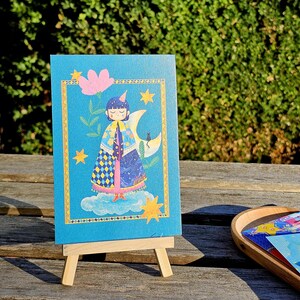 May include: A whimsical illustration on a blue card, featuring a figure in a cape and a party hat, standing on a cloud. The card is framed with a gold border and adorned with stars and a crescent moon. The card is displayed on a small wooden easel.