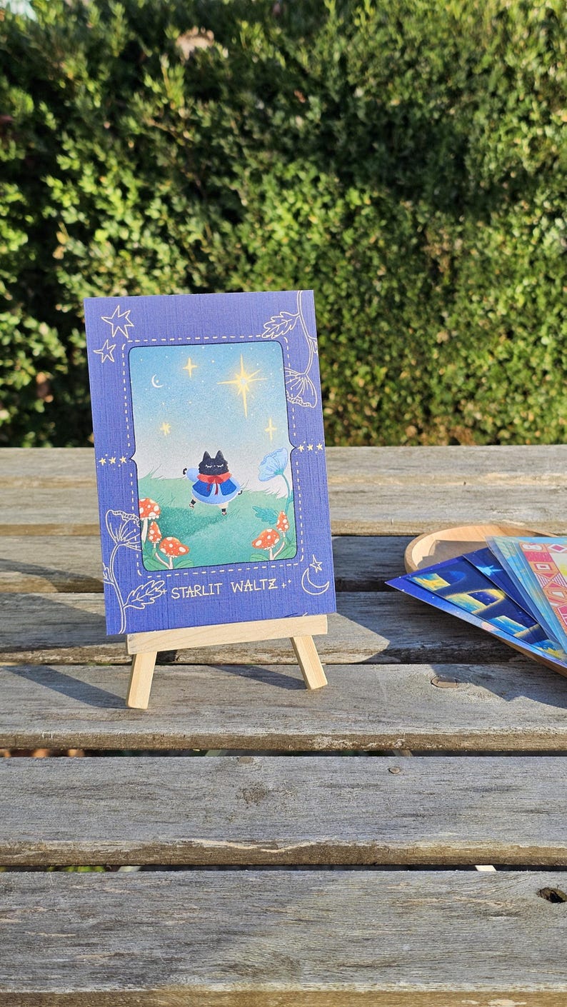 May include: A small, blue framed illustration on a wooden easel. The illustration features a black cat in a blue cape, mushrooms, and the text "STARLIT WALTZ." Additional colorful cards are visible on a wooden surface.