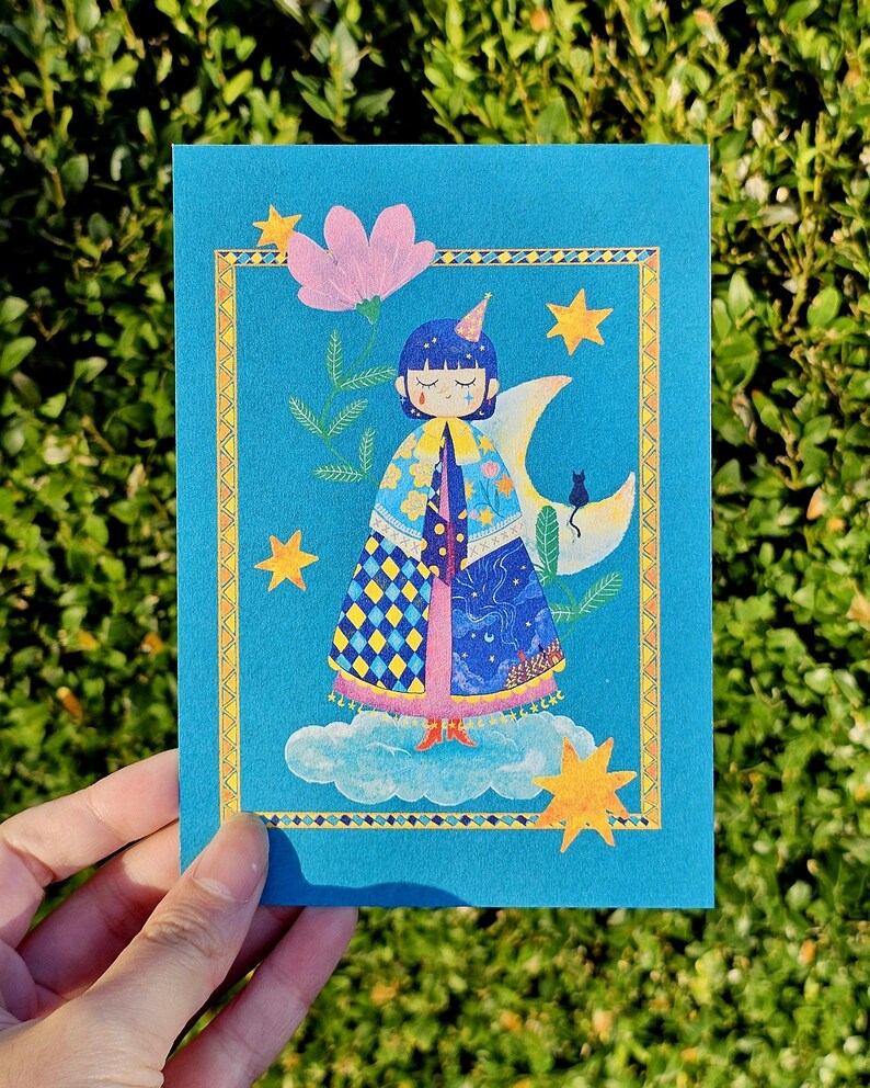 May include: A blue greeting card featuring an illustration of a figure with a unicorn horn, a cape, and a crescent moon. The card has a gold border, stars, a pink flower, and a black cat.