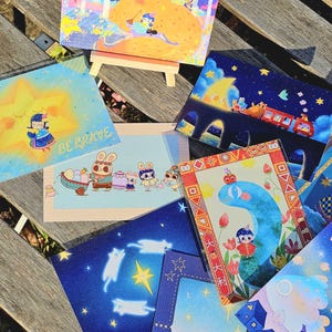 May include: A collection of colorful art prints featuring whimsical illustrations. The prints depict various scenes, including a fox and a child, bunnies with Easter eggs, and a train under a starry sky. Some prints have text that reads "BE BRAVE".