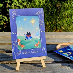 May include: A blue card with a whimsical illustration of a cat in a blue coat dancing under a starry sky. The card is propped on a small wooden easel. The text "STARLIT WALTZ" is visible on the card.