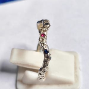 May include: A silver ring with a textured, organic design. The ring features a red and a blue gemstone. The ring is displayed on a white stand against a white background.