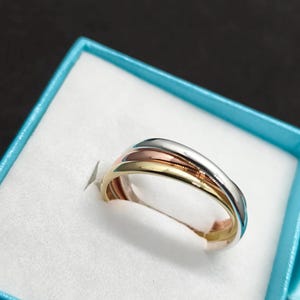 Puede incluir: Un anillo tricolor que presenta tres bandas entrelazadas en tonos oro amarillo, oro rosa y plata. El anillo se muestra descansando en un joyero azul claro con un forro interior blanco, exhibiendo un acabado metálico liso y pulido.
