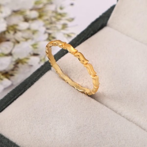 Könnte beinhalten: Ein goldfarbener Ring mit einem sich wiederholenden, strukturierten Design. Der Ring befindet sich vor einem weichen, hellen Hintergrund in einer dunkelgrünen Schmuckschatulle. Das Design des Rings zeigt eine Reihe von geschwungenen, blattartigen Formen.