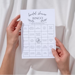 May include: A white Bridal Shower Bingo game card held by hands. The card features a grid with various prompts and the text "Bridal Shower Bingo" at the top. The background is a white fabric.
