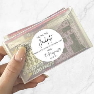 Op de afbeelding: Een hand houdt een stapel loterijloten vast, verpakt in doorzichtig cellofaan. Een witte ronde sticker leest "WE HIT THE Jackpot!" met "The Newlyweds" en de datum 02.05.26. De loten zijn geladen met prijzen van $100.