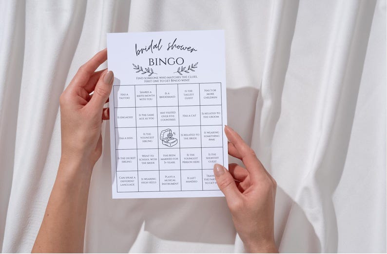 May include: A white Bridal Shower Bingo game card held by hands. The card features a grid with various prompts and the text "Bridal Shower Bingo" at the top. The background is a white fabric.