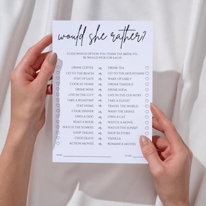 May include: A white card with the text "would she rather?" in a handwritten font. The card lists various choices, such as "drink coffee" or "drink tea", for a bridal shower game. The card is held by two hands.