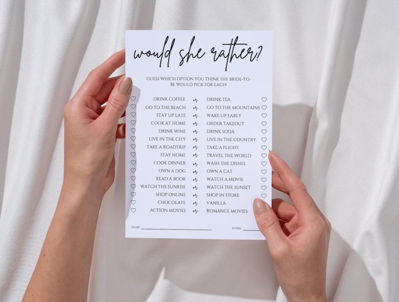 May include: A white card with the text "would she rather?" in a handwritten font. The card lists various choices, such as "drink coffee" or "drink tea", for a bridal shower game. The card is held by two hands.