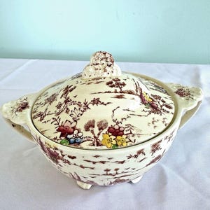 May include: A cream-colored ceramic lidded bowl featuring a brown transferware landscape pattern with hand-painted accents in red, yellow, and blue. The bowl has two side handles, a decorative lid knob, and stands on three small feet.