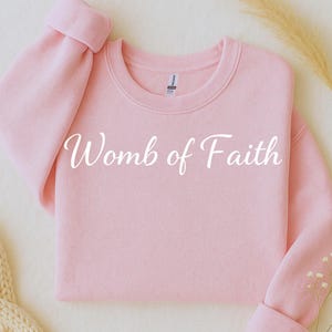 Op de afbeelding: Een lichtroze sweatshirt met de witte tekst "Womb of Faith" in sierlijke letters op de voorkant. De sweatshirt heeft lange mouwen met opgerolde manchetten en een ronde hals. De achtergrond is neutraal.