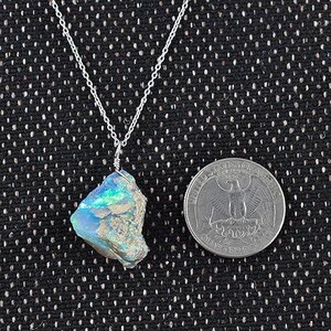 May include: A silver necklace with a raw, iridescent opal pendant. The opal displays blue and green hues. A quarter coin is next to the pendant for size comparison. The necklace chain is delicate.
