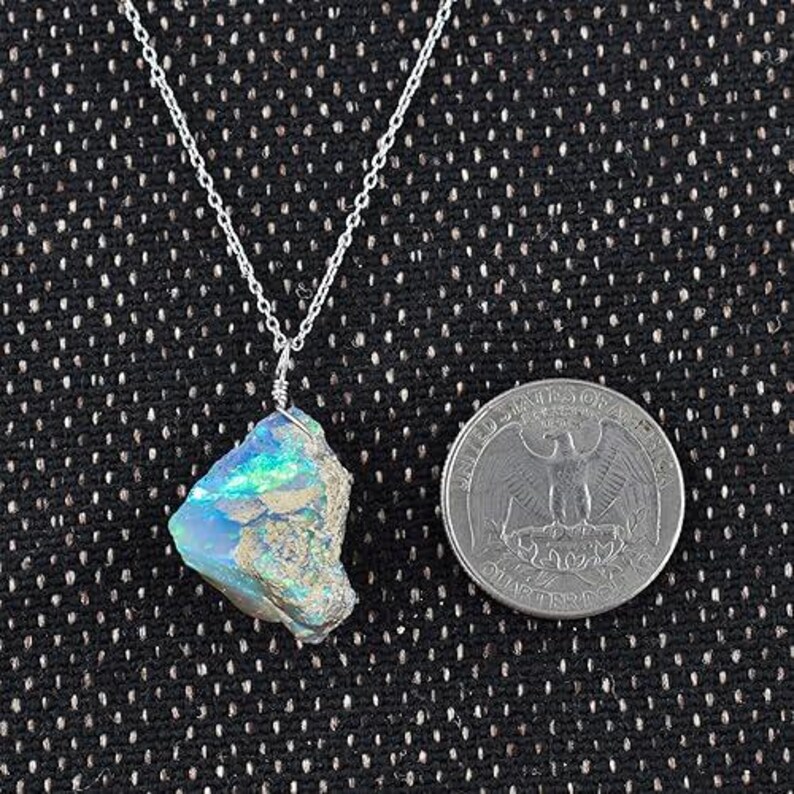 May include: A silver necklace with a raw, iridescent opal pendant. The opal displays blue and green hues. A quarter coin is next to the pendant for size comparison. The necklace chain is delicate.