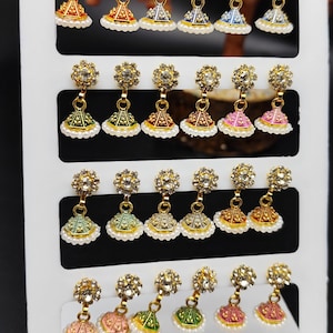 Puede incluir: Una exhibición de pendientes dorados con varios diseños de esmalte de colores, incluyendo rojo, azul, verde y rosa. Cada pendiente presenta una parte superior en forma de flor con colgantes en forma de campana adornados con perlas de imitación. Los pendientes se exhiben en una tarjeta blanca.