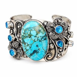 May include: Silver cuff bracelet featuring a large oval turquoise stone at the center. The bracelet is adorned with filigree flowers and smaller blue gemstones. The turquoise stone has a mottled blue and green pattern.