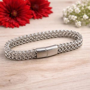 Könnte beinhalten: Ein silberfarbenes Kettenarmband mit geflochtenem Design und rechteckigem Verschluss. Das Armband liegt auf einer hellen Holzoberfläche, im Hintergrund rote und weiße Blumen.