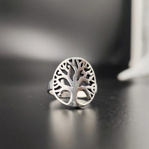 May include: A silver-colored ring featuring a tree of life design. The ring has an oval-shaped face with the tree's branches and trunk cut out. The ring is set against a dark background.