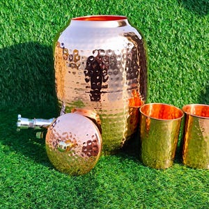 Puede incluir: Un juego de dispensador de agua de cobre martillado con dos tazas a juego. El dispensador tiene un cuerpo grande y redondeado y una tapa. Las tazas y el dispensador tienen una superficie brillante y texturizada. El juego se exhibe sobre hierba verde.