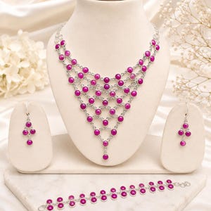 May include: A jewellery set consisting of a multi-strand bib necklace, a pair of drop earrings, and a bracelet. Each piece features round, magenta-coloured cabochon stones set in silver-toned metal frames, arranged in a repeating lattice pattern.