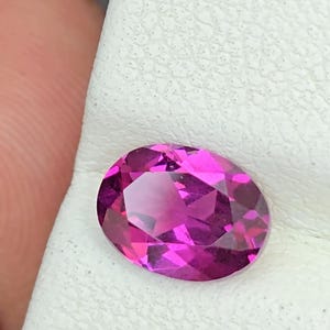 May include: A vibrant, oval-cut, magenta gemstone. The gem is set against a white surface, with a dark background. The stone's facets reflect light, creating a sparkling effect. This gemstone is suitable for jewelry.