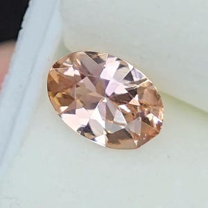 May include: An oval-cut gemstone with a peach color, set against a white background. The gem has multiple facets, reflecting light. The stone is likely a morganite, a gemstone often used in jewelry.