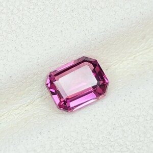 May include: A faceted, rectangular-cut gemstone with a vibrant pink hue. The gem has a clear, transparent center and a darker pink border. The stone is set against a textured, off-white surface.