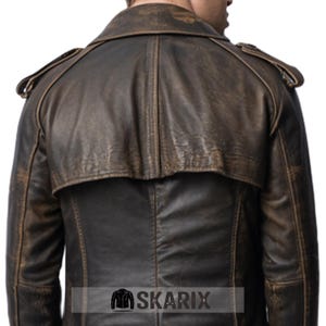 May include: A distressed brown leather jacket with a classic trench coat design. The jacket features a notched collar, shoulder epaulets, and a back cape. The leather has a worn, vintage appearance, with visible texture and color variations. The brand name "SKARIX" is at the bottom.