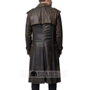 May include: A long, brown leather coat with a distressed finish. The coat features a double-breasted design, epaulettes on the shoulders, and a long, flowing skirt. The back of the coat has a vent at the bottom. The Skarix logo is visible at the bottom.