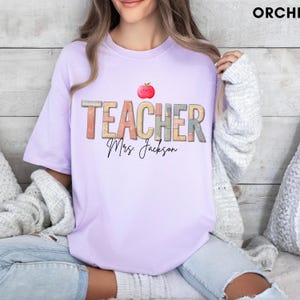 Puede incluir: Una camiseta de manga corta de color morado claro con la palabra TEACHER en una fuente multicolor con patrón de lomos de libros. Sobre el texto hay un pequeño gráfico de una manzana roja sonriente y bajo el texto el nombre Mrs. Jackson en negro.