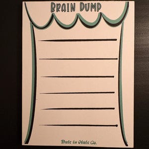 May include: A cream-colored Brain Dump notepad with black horizontal lines for writing. The top of the notepad has a scalloped design with teal and black accents. The words "Brain Dump" are printed at the top, and "Pals in Hats Co." is printed at the bottom.