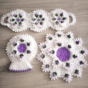 Puede incluir: Un conjunto de tres artículos de ganchillo hechos a mano con hilo blanco y detalles florales en morado y verde. La colección incluye una pieza larga de tres secciones con asas, una pieza redondeada con base y un tapete circular grande.