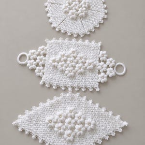 Puede incluir: Tres piezas de ganchillo blancas con intrincados patrones florales y pequeñas cuentas perladas. El conjunto incluye un tapete circular, una pieza rectangular con anillos laterales y una pieza en forma de diamante, todas con bordes festoneados.