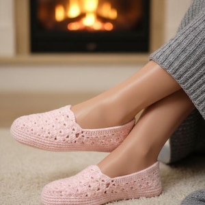 Może przedstawiać: Para jasnoróżowych kapci zrobionych na szydełku. Kapcie mają ażurowy wzór i zaokrąglony nosek. Obraz przedstawia kapcie na stopach osoby, z kominkiem w tle. Kapcie wydają się miękkie i wygodne.