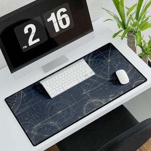 May include: A desk setup with a dark blue mouse mat featuring a gold blueprint design. A white keyboard and mouse are on the mat. A computer monitor displays the time, and a potted plant is in the background. The mat is approximately 80 cm long.