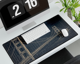 May include: A black rectangular mousepad featuring a blueprint-style illustration of the Golden Gate Bridge in tan and white. A white keyboard and mouse sit on the pad, with a computer monitor and a plant in the background.