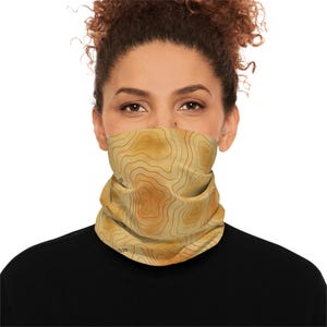 May include: A tan and brown neck gaiter with a topographic map design. The gaiter is wrapped around the lower face and neck. The map design features contour lines and the number "1500". The person is wearing a black shirt.