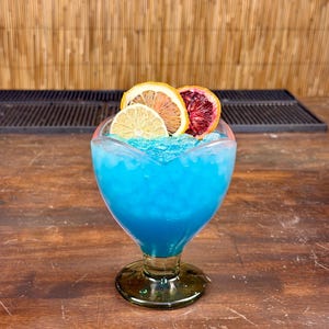 May include: A vibrant blue cocktail in a heart-shaped glass, garnished with dried citrus slices. The drink sits on a wooden surface, with a bamboo backdrop. The glass has a green stem and base, and the drink is filled with crushed ice.