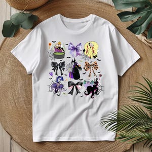 K&ouml;nnte beinhalten: Wei&szlig;es T-Shirt mit einem Halloween-Motiv mit bunten Schleifen. Die Schleifen zeigen verschiedene Charaktere und Elemente, darunter eine Hexe, eine K&ouml;nigin und einen Oktopus. Das Design enth&auml;lt Spinnennetze, Flederm&auml;use und andere gruselige Details.