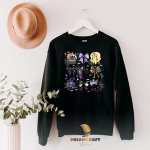 K&ouml;nnte beinhalten: Schwarzes Sweatshirt mit einem Halloween-Motiv. Das Design umfasst Hexen, Schleifen, Spinnennetze und einen Kessel. Das Sweatshirt h&auml;ngt an einem wei&szlig;en Kleiderb&uuml;gel vor einer wei&szlig;en Wand. Das Bild enth&auml;lt auch einen Hut und eine Pflanze.