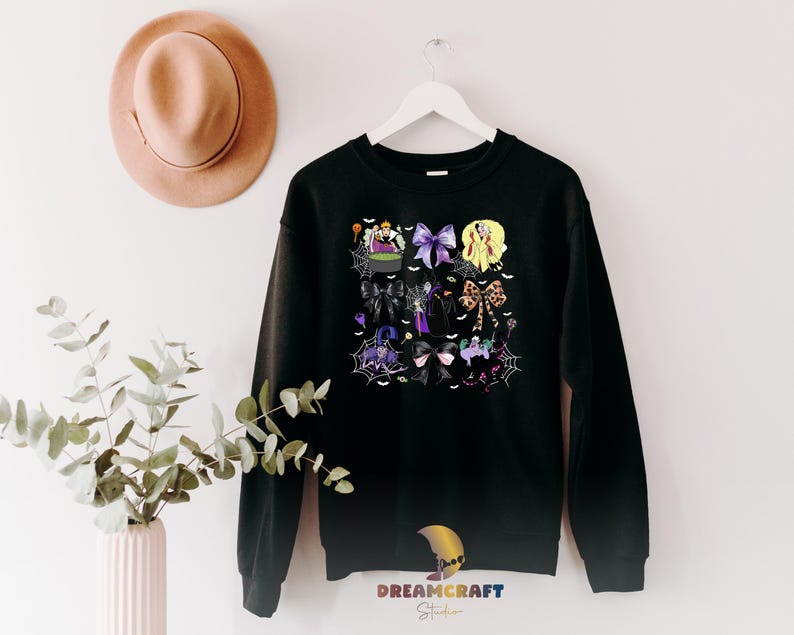 K&ouml;nnte beinhalten: Schwarzes Sweatshirt mit einem Halloween-Motiv. Das Design umfasst Hexen, Schleifen, Spinnennetze und einen Kessel. Das Sweatshirt h&auml;ngt an einem wei&szlig;en Kleiderb&uuml;gel vor einer wei&szlig;en Wand. Das Bild enth&auml;lt auch einen Hut und eine Pflanze.