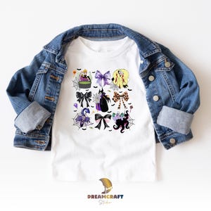K&ouml;nnte beinhalten: Wei&szlig;es T-Shirt mit einem farbenfrohen Halloween-Motiv mit Schleifen, Hexen und Spinnennetzen. Das Shirt wird von einer blauen Jeansjacke bedeckt. Das Dreamcraft Studio-Logo ist unten sichtbar.