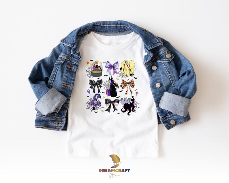 K&ouml;nnte beinhalten: Wei&szlig;es T-Shirt mit einem farbenfrohen Halloween-Motiv mit Schleifen, Hexen und Spinnennetzen. Das Shirt wird von einer blauen Jeansjacke bedeckt. Das Dreamcraft Studio-Logo ist unten sichtbar.