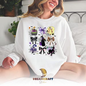 K&ouml;nnte beinhalten: Wei&szlig;es Sweatshirt mit einem farbenfrohen Aufdruck von Halloween-Schleifen und -Figuren. Das Design umfasst lila, schwarze und gelbe Schleifen, Spinnweben und Zeichentrickfiguren. Das Sweatshirt hat lange &Auml;rmel und einen Rundhalsausschnitt.