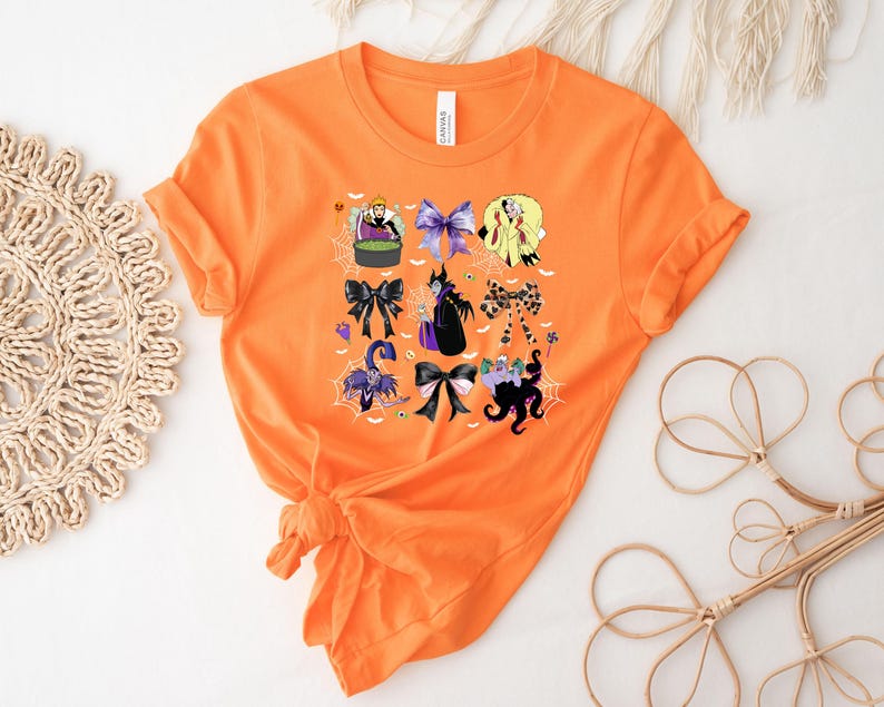 K&ouml;nnte beinhalten: Orangefarbenes T-Shirt mit einem Halloween-Motiv. Das Design zeigt verschiedene Schleifen und Cartoon-B&ouml;sewichte, mit Spinnweben und einem Kessel. Das T-Shirt besteht aus einem weichen Material und hat einen Rundhalsausschnitt.
