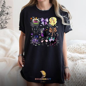 K&ouml;nnte beinhalten: Schwarzes T-Shirt mit einem farbenfrohen Cartoon-Design von Schleifen und Figuren. Das Design umfasst lila, gelbe und schwarze Schleifen, Spinnweben und Illustrationen zum Thema Hexen. Das T-Shirt hat unten den Text "DREAMCRAFT Studio".