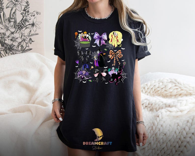 K&ouml;nnte beinhalten: Schwarzes T-Shirt mit einem farbenfrohen Cartoon-Design von Schleifen und Figuren. Das Design umfasst lila, gelbe und schwarze Schleifen, Spinnweben und Illustrationen zum Thema Hexen. Das T-Shirt hat unten den Text "DREAMCRAFT Studio".
