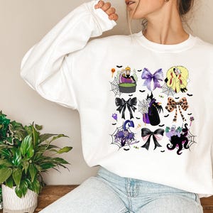 K&ouml;nnte beinhalten: Wei&szlig;es Sweatshirt mit einem farbenfrohen Grafikdesign mit Halloween-Motiven, darunter Schleifen und Charaktere. Das Design zeigt eine Hexe, eine K&ouml;nigin und andere gruselige Elemente. Das Design ist auf der Vorderseite des lang&auml;rmeligen Oberteils aufgedruckt.